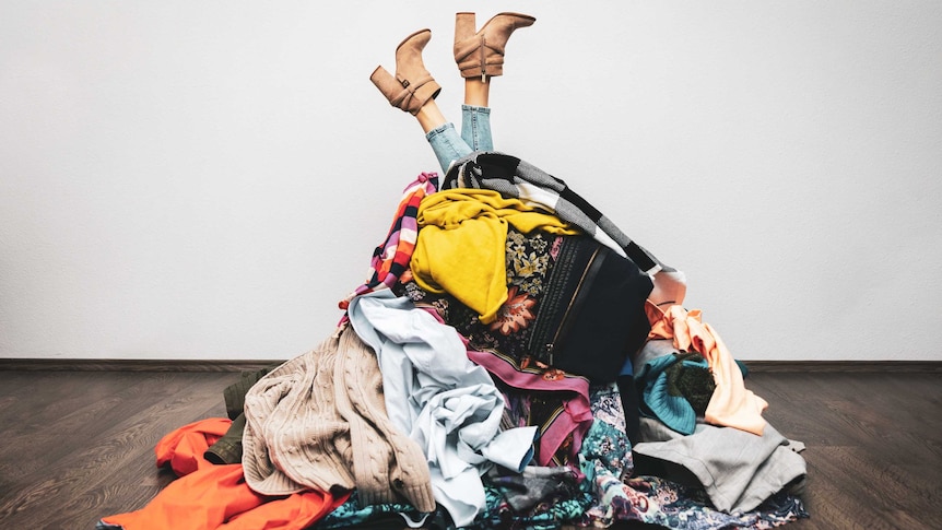 A large mound of clothes are viewed on a wooden floor with a person's legs poking out from the top. 