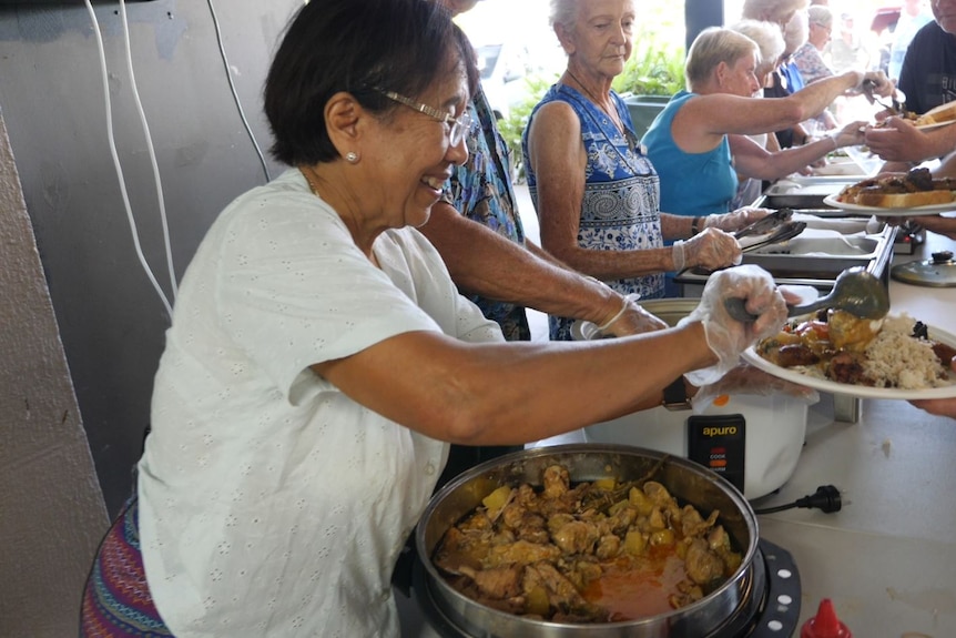 Woman wearing a white shirt serves chicken curry onto a plate held by another person.