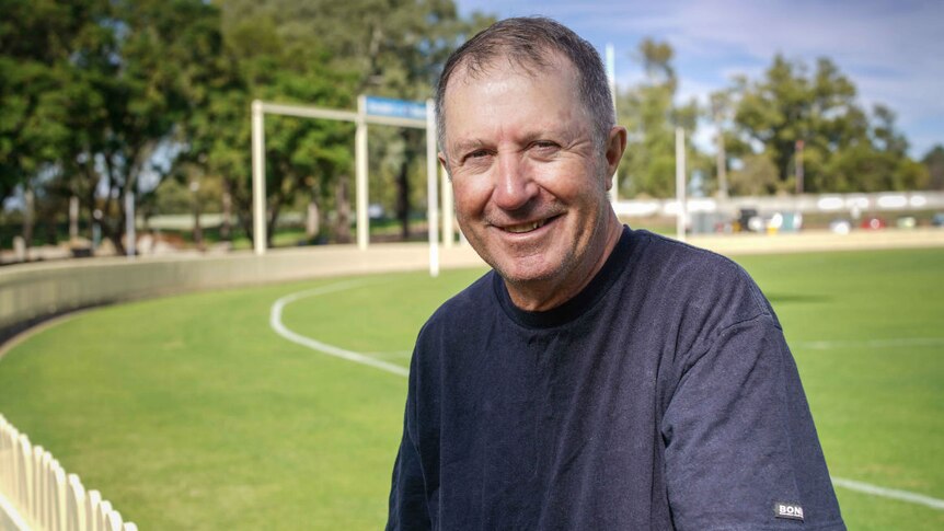 A older man with short hair, wearing a blue top, standing in front of a sports ground.