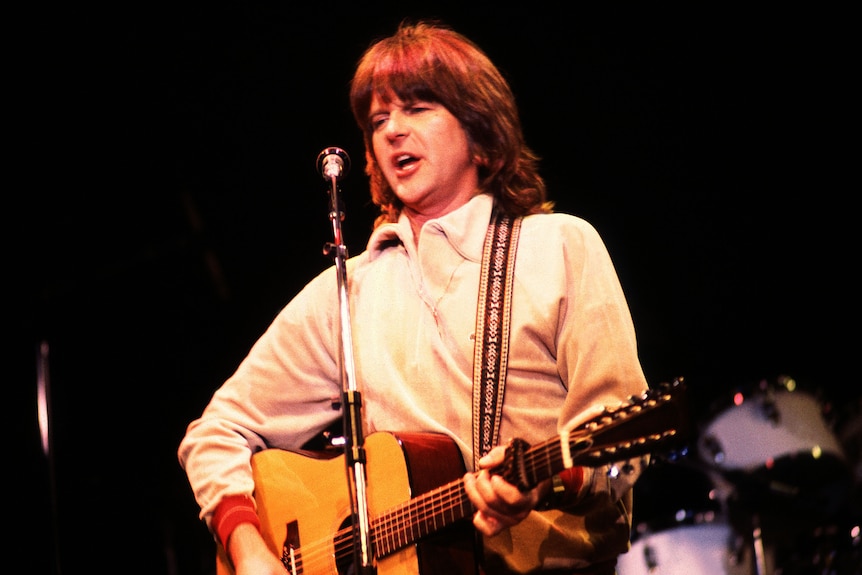 A man with long wearing a white sweater playing a guitar and singing into a microphone with a dark background