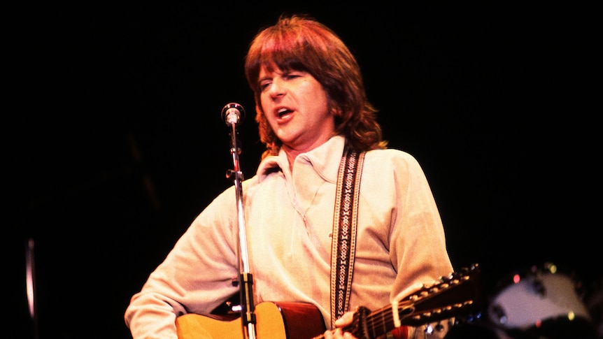 A man with long wearing a white sweater playing a guitar and singing into a microphone with a dark background