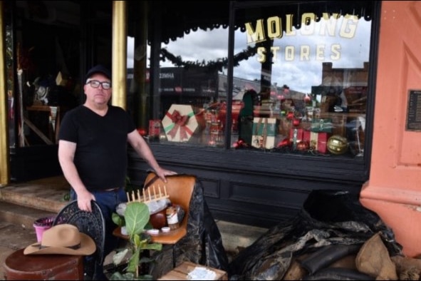 A middle-aged man in black tee and black cap standing next to a flooded shop. 