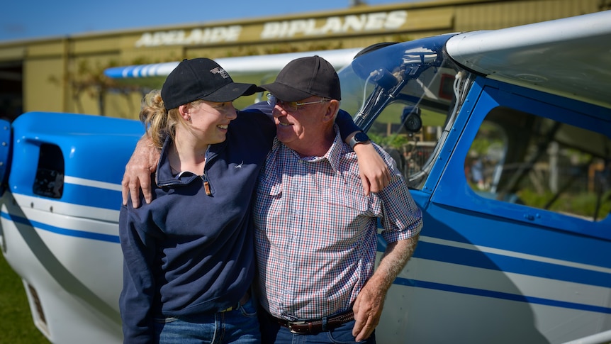 A teen girl with her arm around her grandfather looking at each other, standing in front of a light aircraft