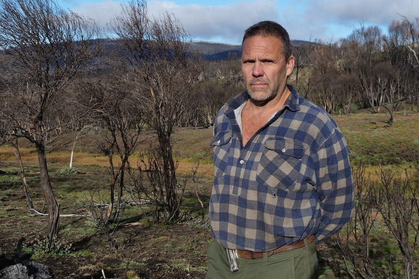 Een man met bruin haar en stoppels die een flannelette shirt draagt en in een uitgebrand deel van Kosciuszko National Park staat.