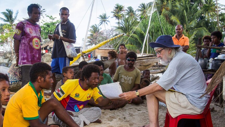 Elderly man interviewing island villagers on beach in front of sailing canoes.