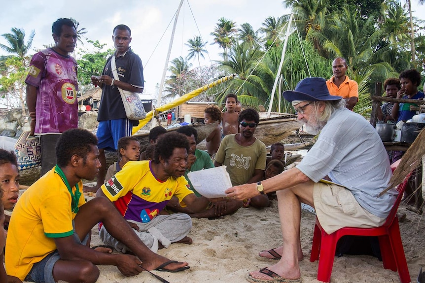 Elderly man interviewing island villagers on beach in front of sailing canoes.