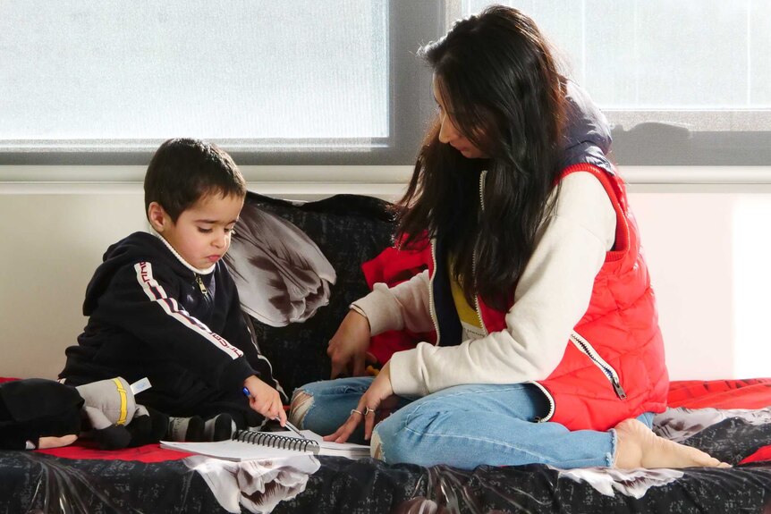 A young boy sits on a bed with his mother, holding a pen and pointing at some paper.