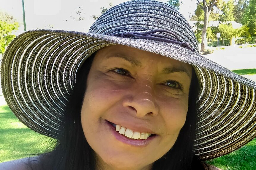Woman wearing wide brim hat smiles looking at camera with park in background.