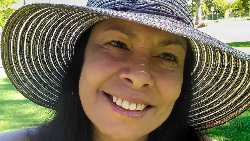 Woman wearing wide brim hat smiles looking at camera with park in background.