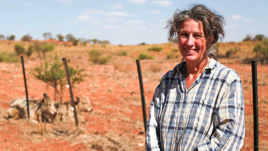 A woman in a checked shirt stands next to a wire fence on red dirt