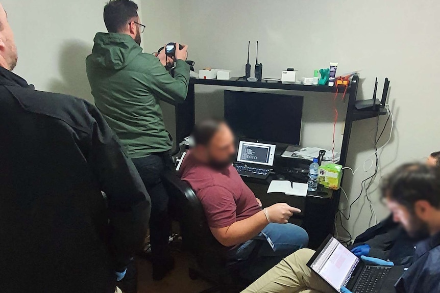 There are five men in the photo, including one sitting at a desk. Officers are taking photos and searching computers.
