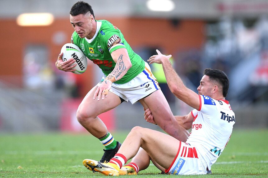 A Canberra Raiders NRL player holds the ball with his right hand as a Dragons opponent tackles him around his left leg.