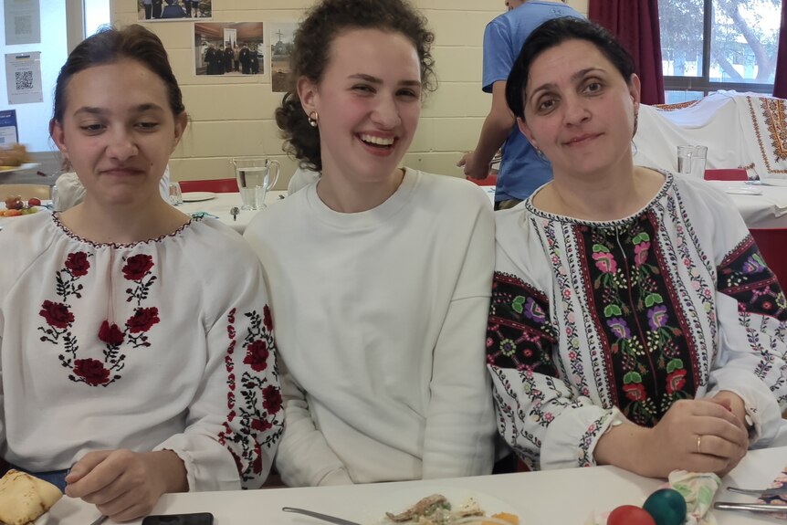 Three women in white patterned clothing at a table smiling.