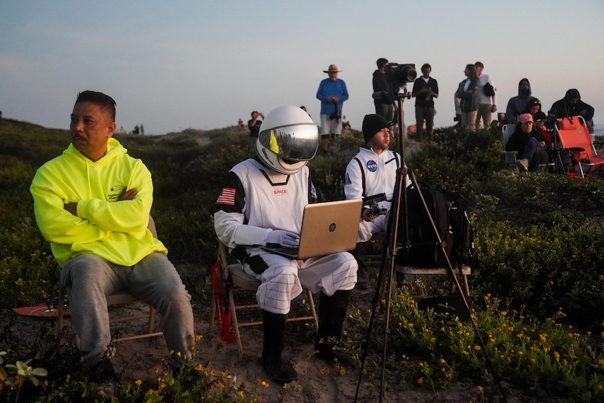 A person in an astronaut costume sits with a computer and camera on a hill 