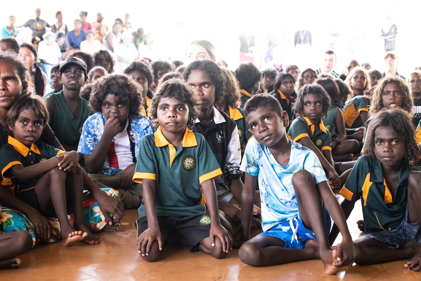 A group of indigenous children watch off-camera at a stage outside of the image frame.