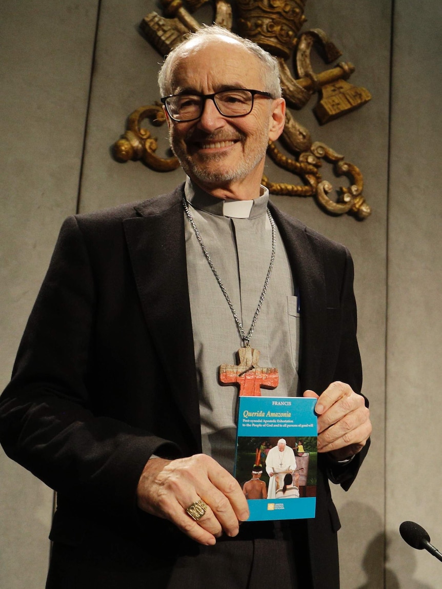 An older man with glasses and Catholic collar smiles and holds a small book.