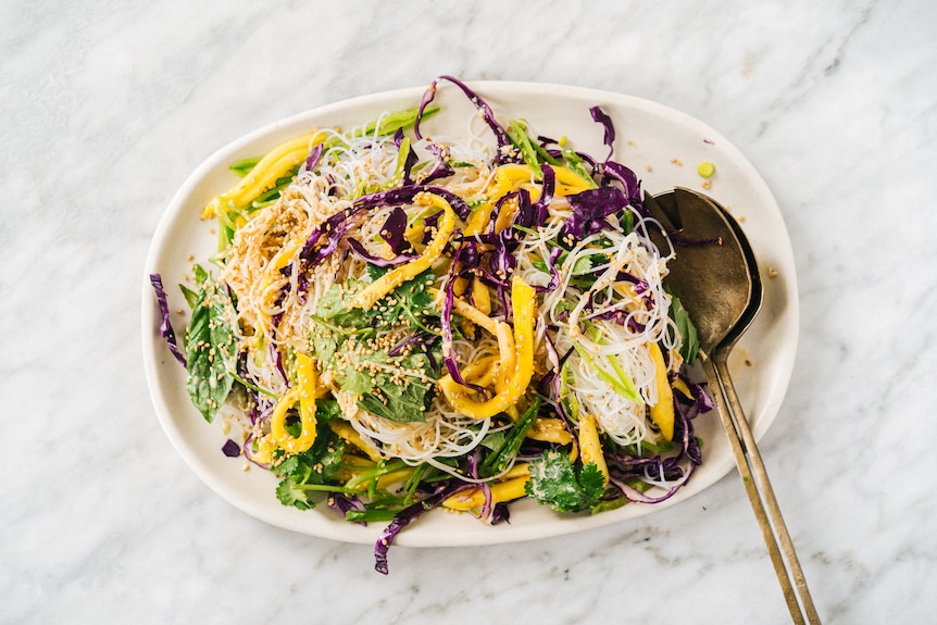 A serving bowl of colourful noodle salad featuring vermicelli and slivers of yellow, purple and green ruit and veg.