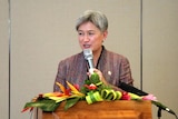 A woman in a jacket smiles as she speaks into a mic that's rigged to a lectern adorned with flowers. 