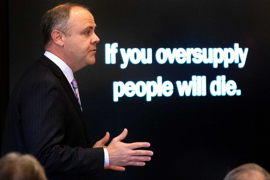 State attorney Brad Beckworth speaks with a screen which reads, "If you oversupply people will die", behind him.