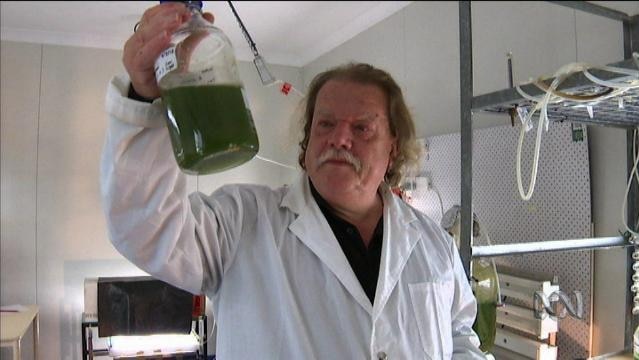 A man in a white coat holds up a jar of green liquid