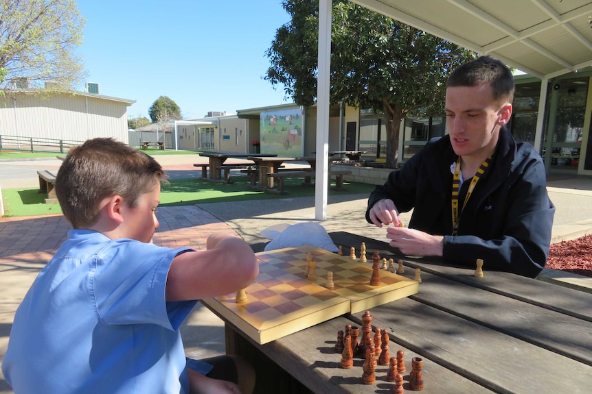A young man plays chess on a wooden bench seat with a student on school grounds.