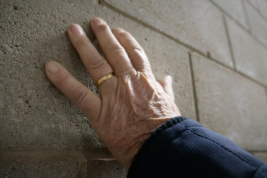 A close up image of a senior man's hand against a brick wall. It has worked hard and is wrinkled. He wears a gold wedding band.