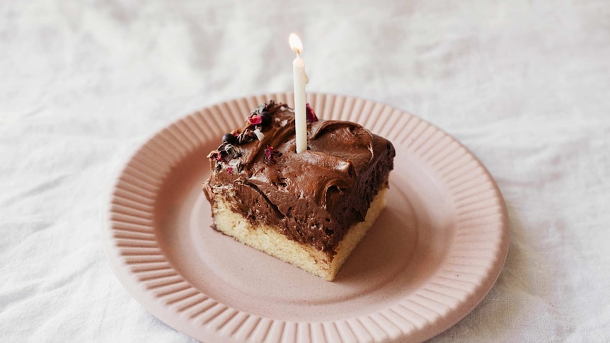 A slice of vanilla cake with thick chocolate hazelnut icing, edible flowers and a birthday candle, ready for a celebration.