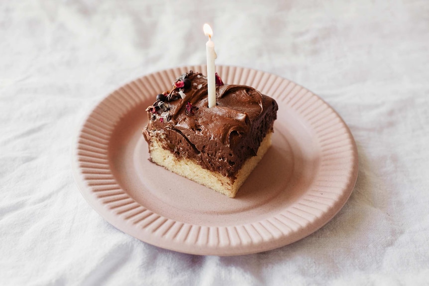 A slice of vanilla cake with thick chocolate hazelnut icing, edible flowers and a birthday candle, ready for a celebration.