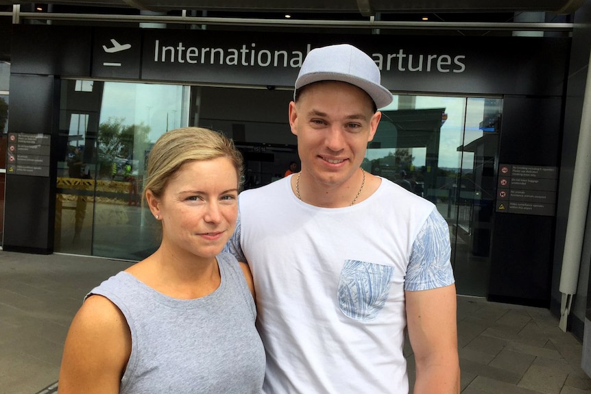 Head and shoulders shot of Emma and Nick outside a sign reading International Departures at Perth Airport.