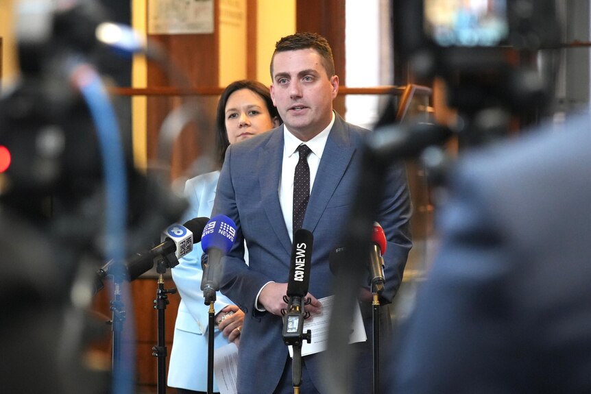 Man in suit with black tie and brown hair addresses media