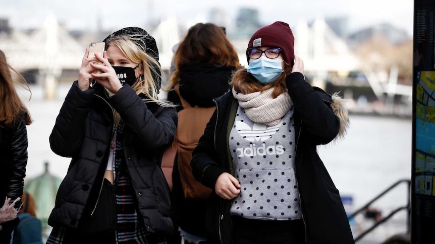 Two women stand at a monument in London wearing protective face masks. One is taking a photo on her phone