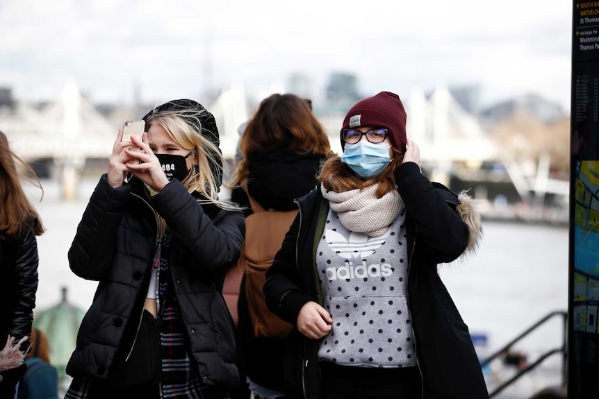 Two women stand at a monument in London wearing protective face masks. One is taking a photo on her phone