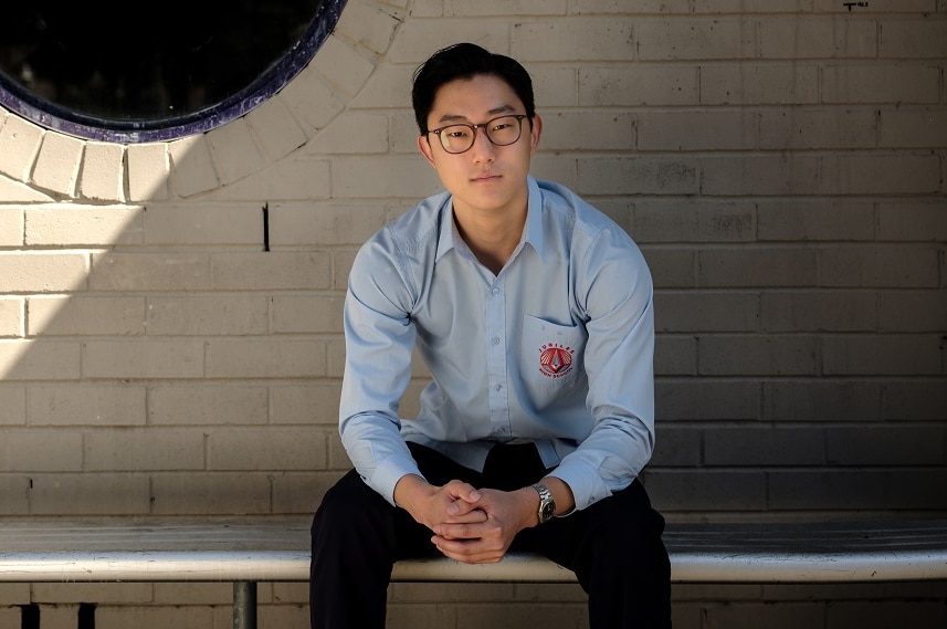 A man wearing glasses sitting on a school bench in blue school uniform in front of a wall with a black circle window