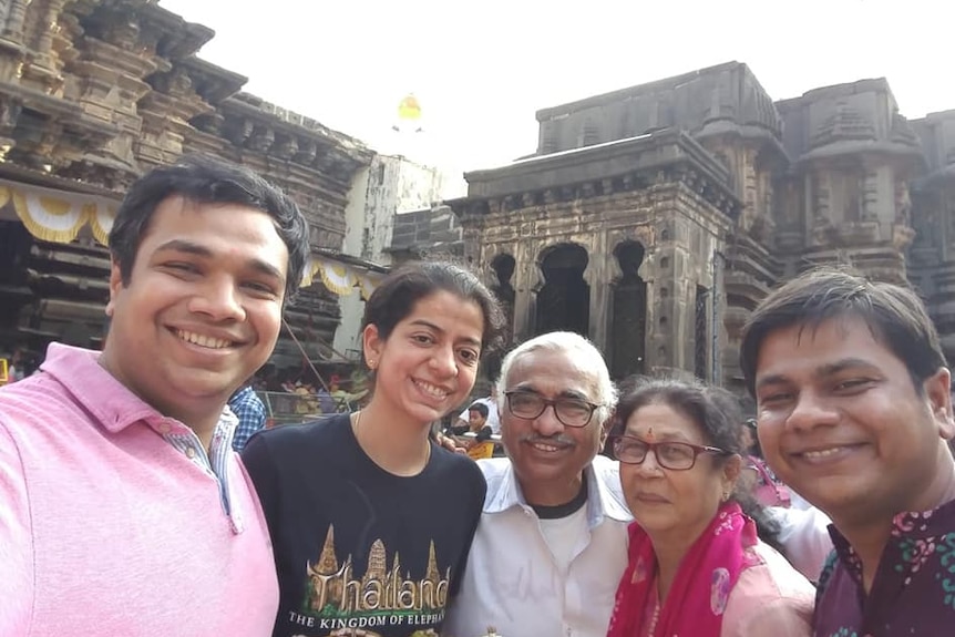 Three young people, stand with an older couple in front of an ancient Hindu temple, wall carvings visible. All smile.