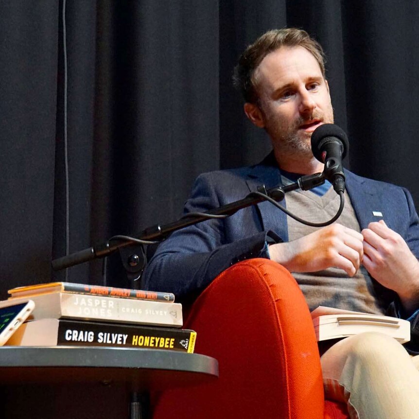 Craig Silvey with a beard talking into a microphone next to a table with his books on it.