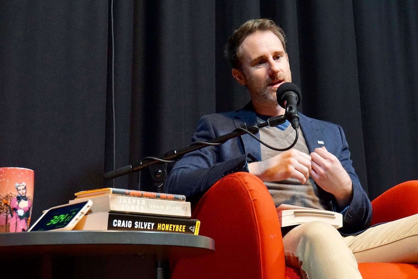 Craig Silvey with a beard talking into a microphone next to a table with his books on it.