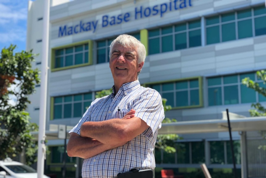 A man stands with his arms crossed in front of a hospital