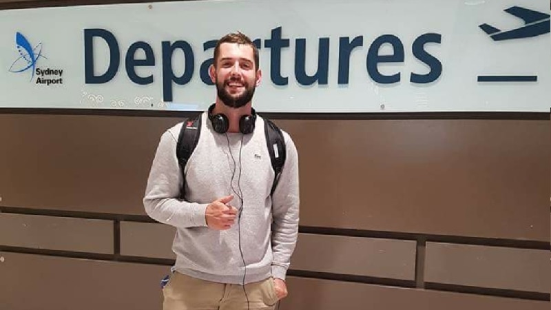 Man smiling with headphones on standing in front of a Sydney Airport departure sign.