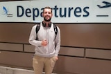 Man smiling with headphones on standing in front of a Sydney Airport departure sign.