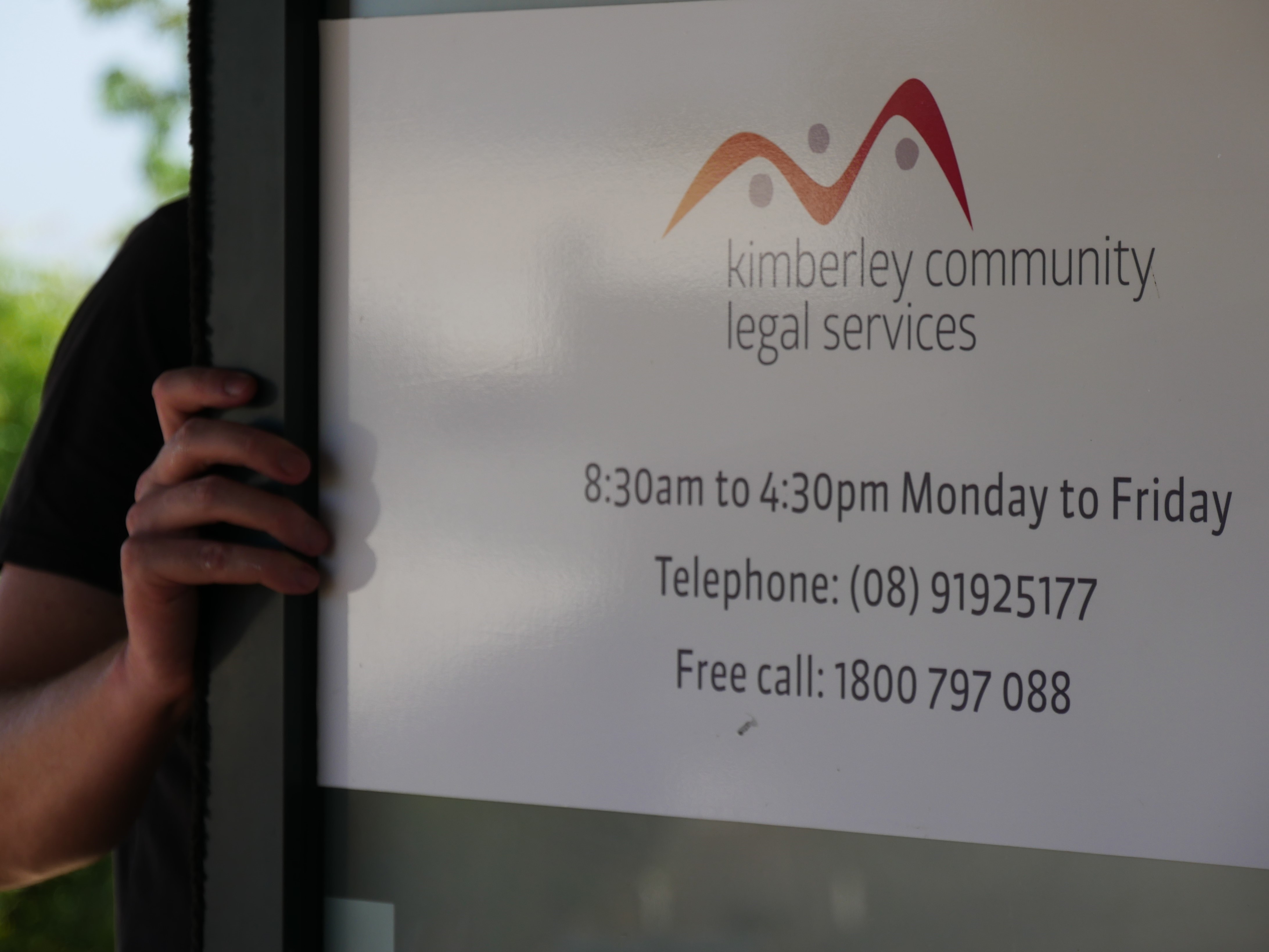 A hand pushing a white door open which has a sign that says Kimberley Community Legal Services.