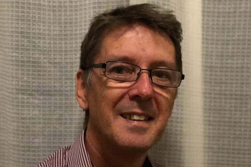 Medium close up of a man with glasses wearing a red and white striped shirt sitting in front of a white curtain.