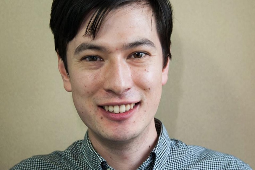 A headshot of a smiling man wearing a checked shirt.