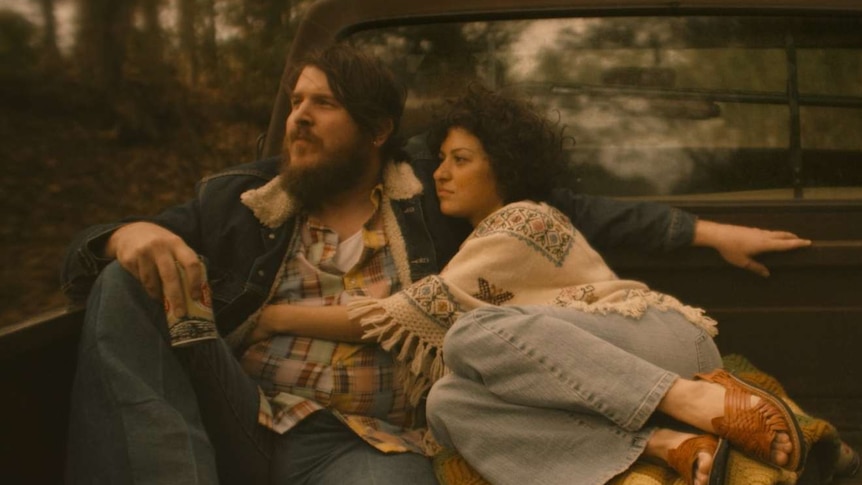 Man and woman wearing 70s Texas country garb embracing in back of flat bed truck driving through countryside.
