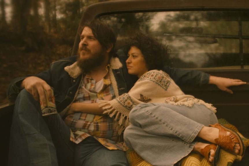 Man and woman wearing 70s Texas country garb embracing in back of flat bed truck driving through countryside.