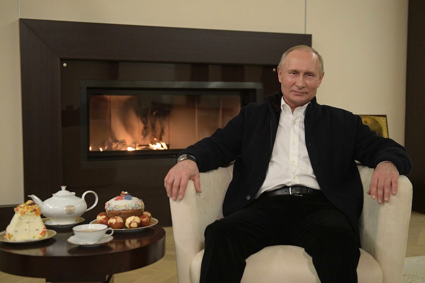 Vladimir Putin sits in front of a fire with a pot of tea, cake and easter eggs beside him.