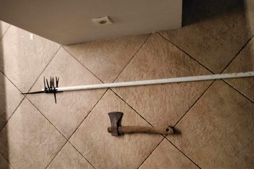 A broom with a knife strapped to it and a tomohawk lie on a floor.