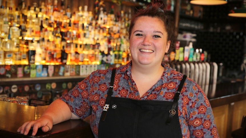 A smiling woman in a colourful shirt and black apron rests her arm on a bar, in front of a backlit row of drinks.