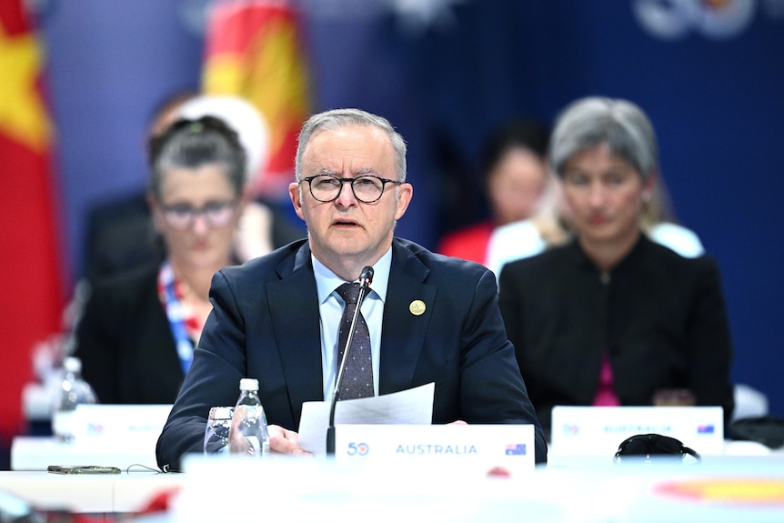 A wide shot of a man in a suit and tie sitting and speaking into a microphone, with people sitting and flags standing behind