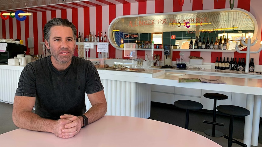 A man in a black shirt with grey stubble sits at a cafe table.