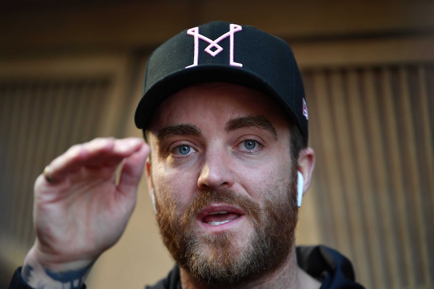 Paul O'Shaughnessy is seen in a headshot and Apple AirPods in his ears, with a cap on his head as he gesticulates.